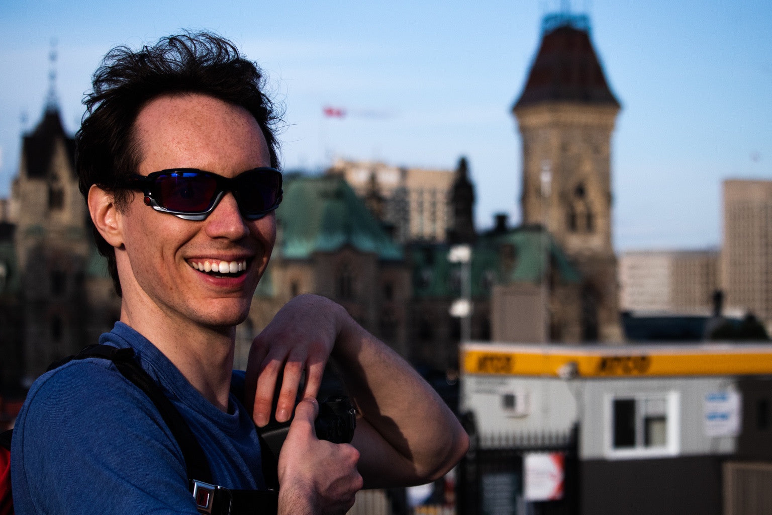 Un photographe souriant tenant un appareil photo, avec des édifices d’Ottawa en arrière-plan