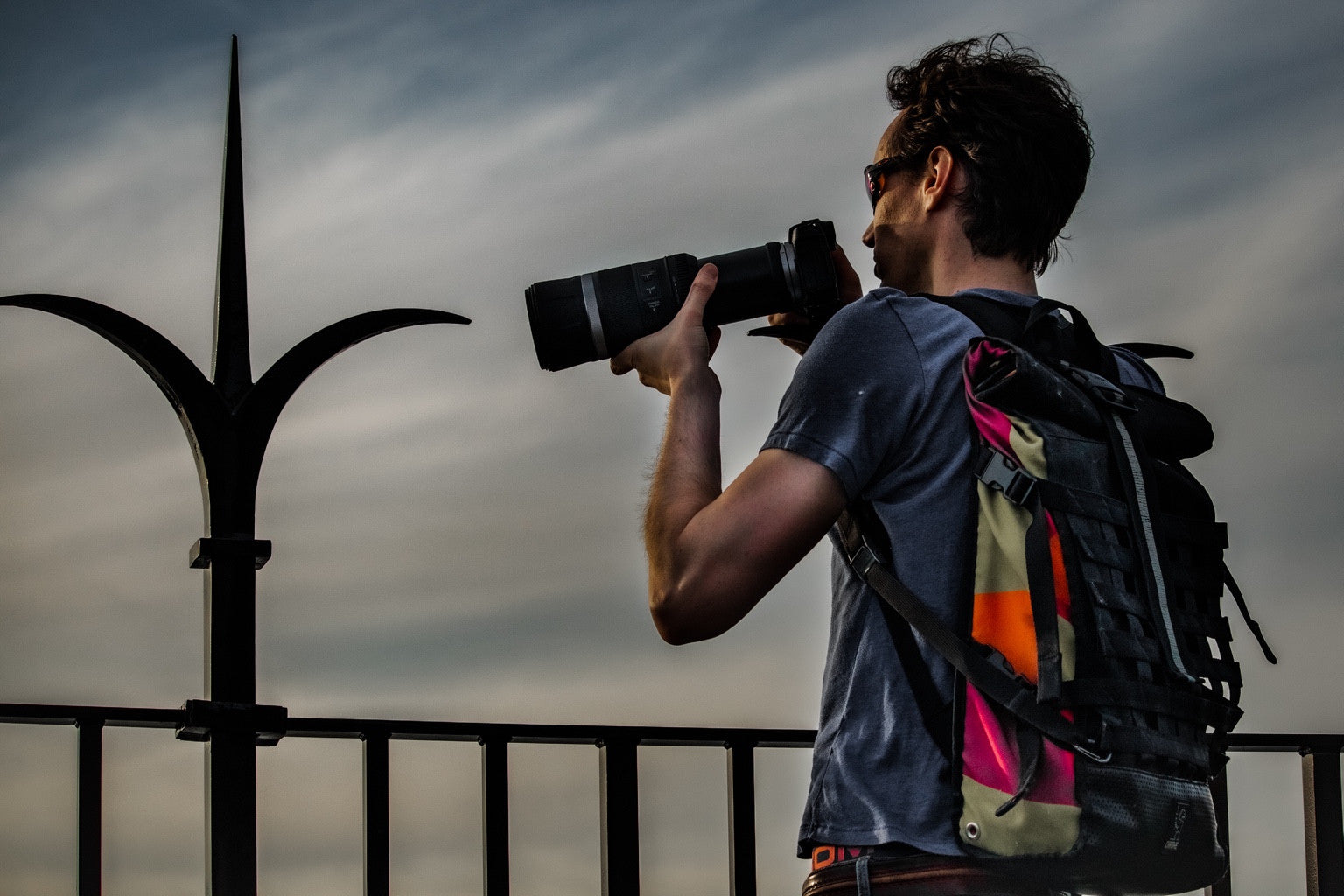 A photographer with a telephoto camera and colourful backpack beside an iron fence